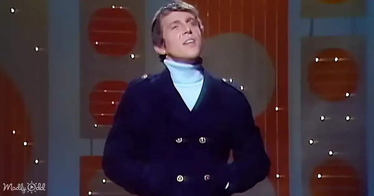 A man in a dark coat and white turtleneck stands against a backdrop with circular patterns and lights.