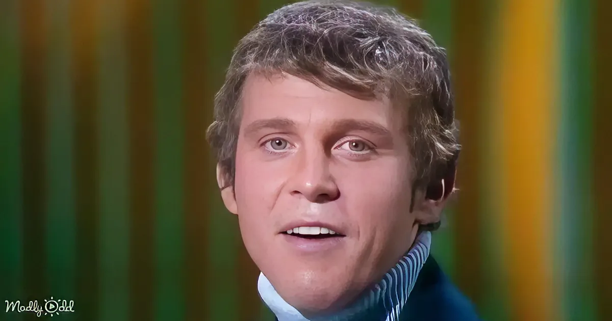 A man with short, curly hair wearing a blue turtleneck sweater, set against a colorful striped background.