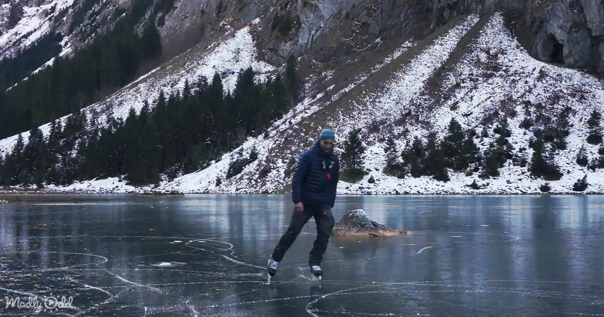 A man ice skating on a frozen lake surrounded by snowy mountains and evergreen trees.