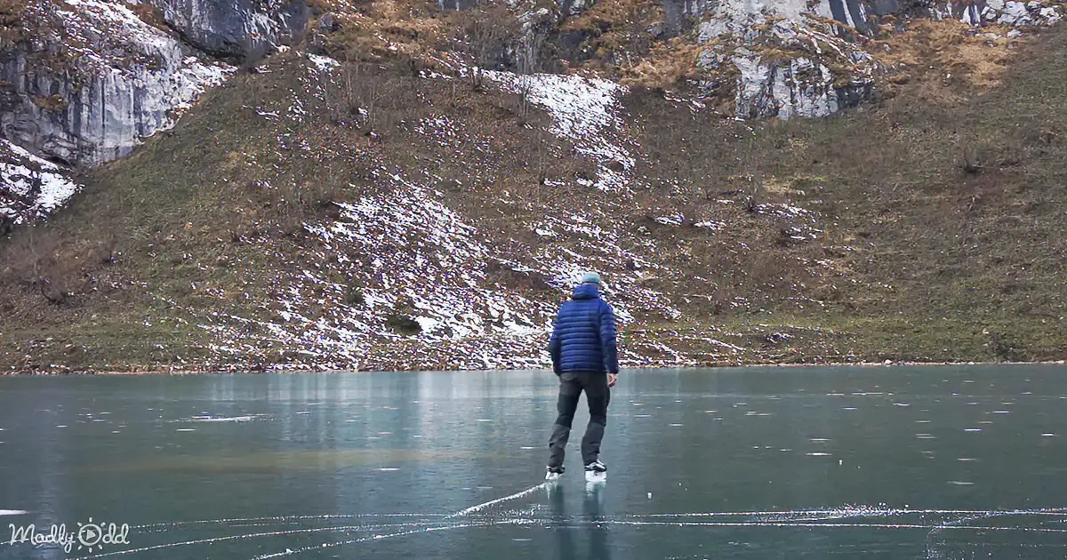 A person in a blue jacket stands on thin ice, facing a snowy, grassy hillside.