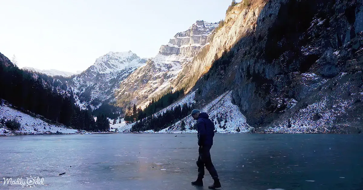 A person walks on a frozen lake surrounded by snow-covered mountains and evergreen trees.