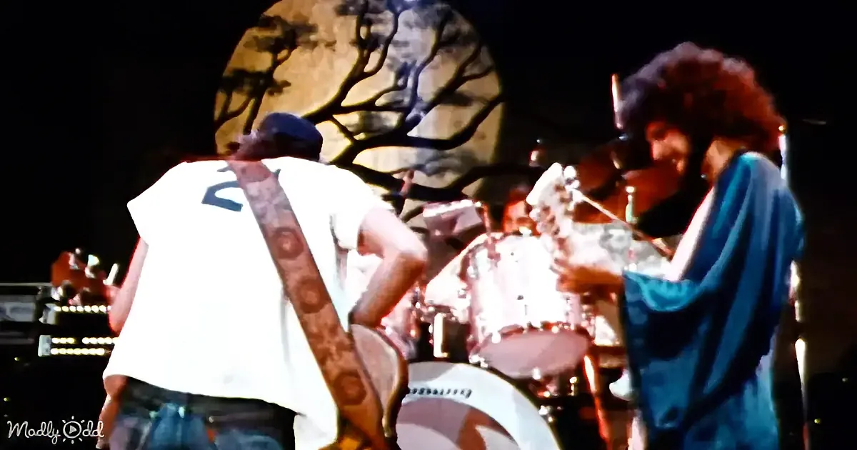 A guitarist in a white shirt and blue jeans plays on stage with a drummer in a blue shirt.