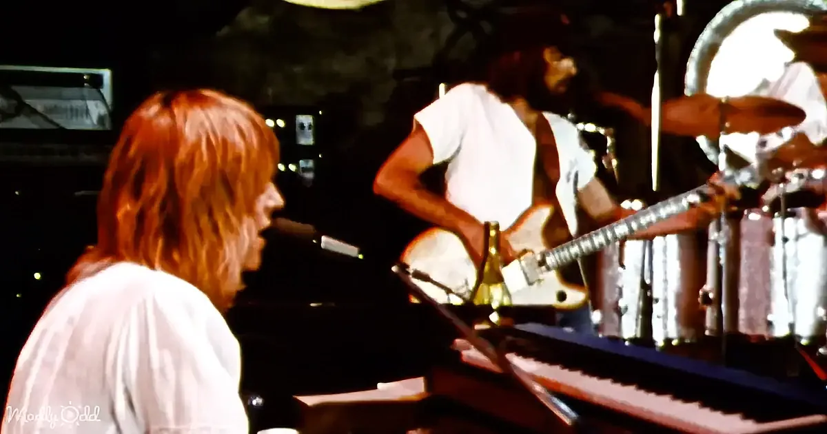 A musician with long hair plays piano, another plays guitar in a dimly lit stage setting.