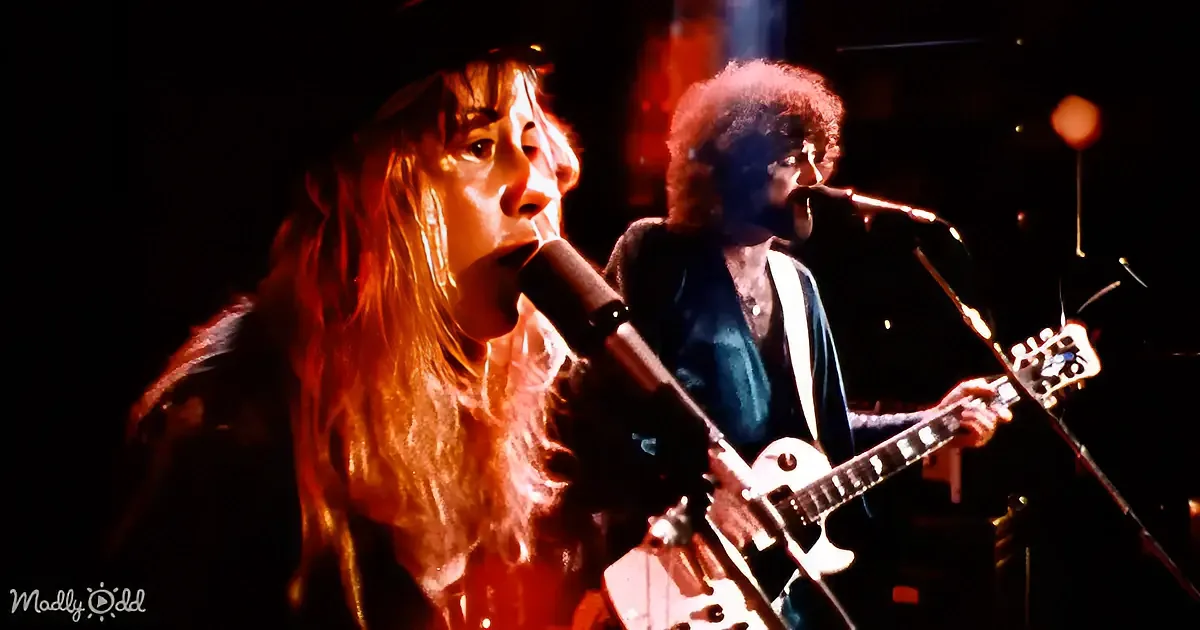 A musician with long hair sings into a microphone, illuminated by stage lights, with another musician playing guitar in...