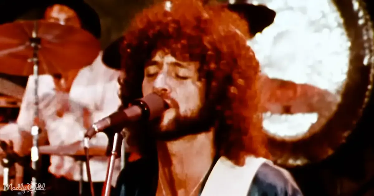 A person with curly hair sings into a microphone on stage, surrounded by musical instruments.