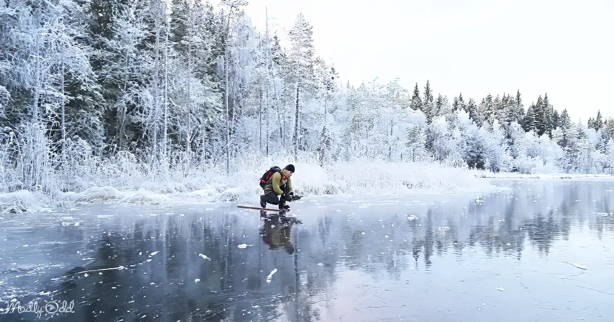 A person in winter gear kneels on a frozen lake, surrounded by snow-covered trees.