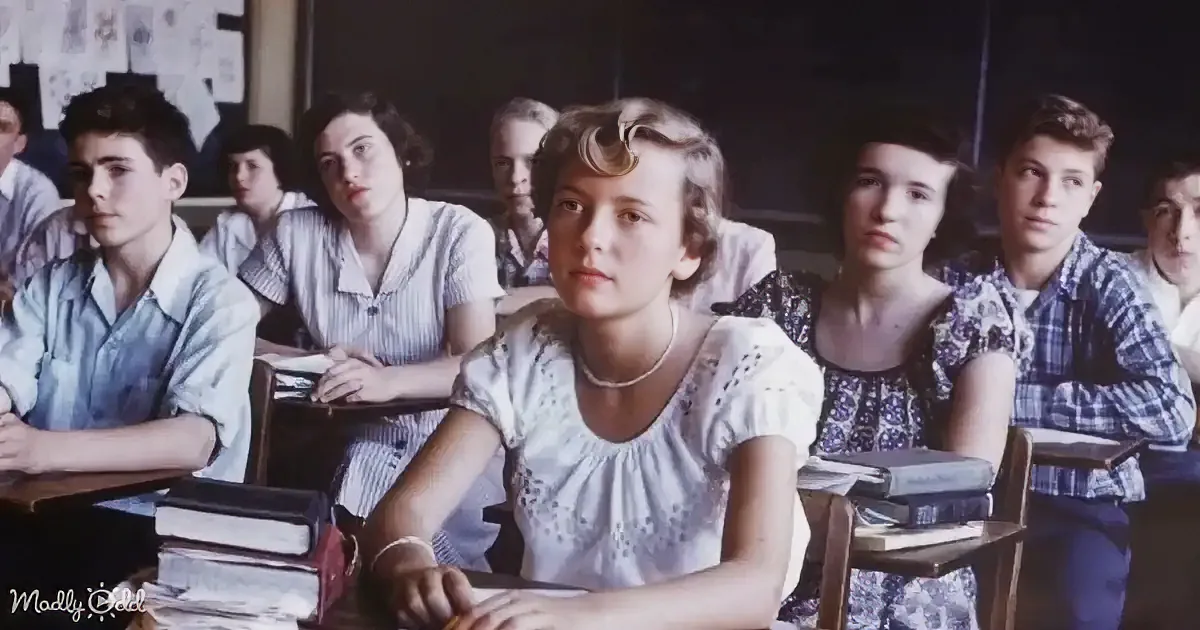 A young woman in a white blouse sits at a desk in a classroom, surrounded by other students.