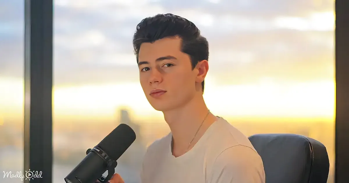 A person in a white shirt holds a microphone, seated against a backdrop of a sunset through a window.