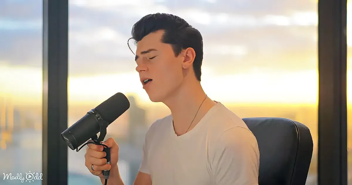 A person in a white shirt sings into a microphone, set against a sunset-lit window backdrop.