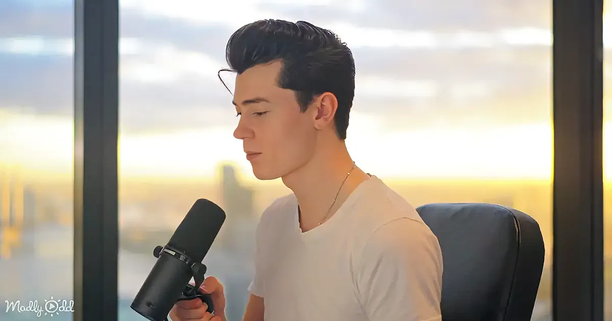 A person in a white shirt sits in front of a microphone, holding it, with a cityscape and sunset in the background.