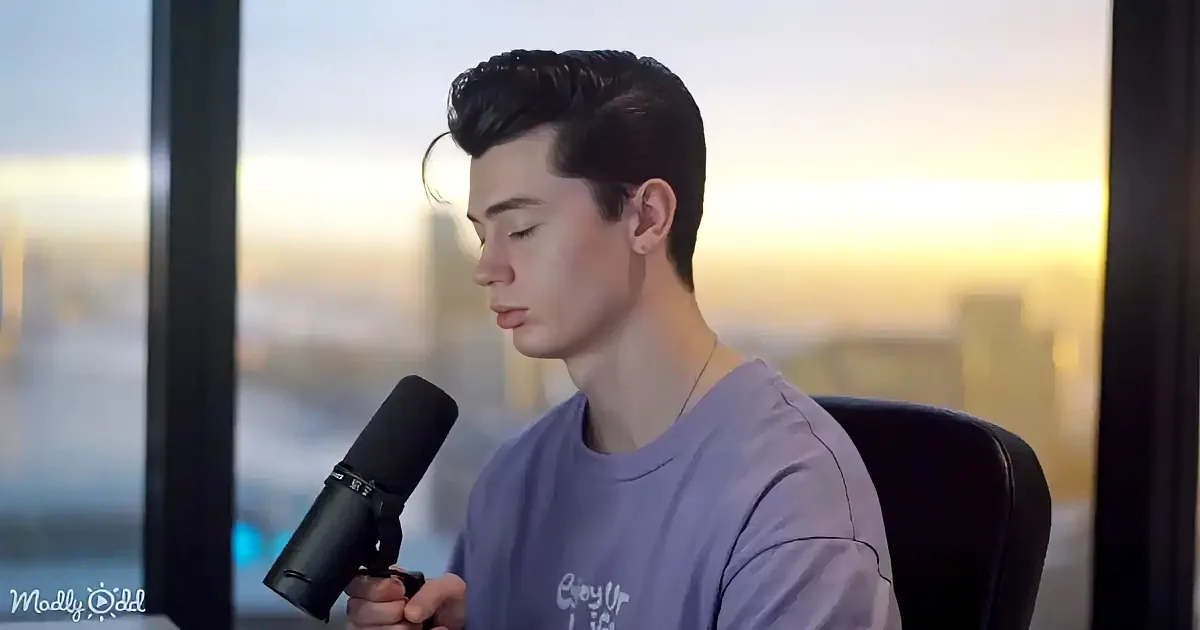 A person in a purple shirt holds a microphone, seated in front of a window with a cityscape view.