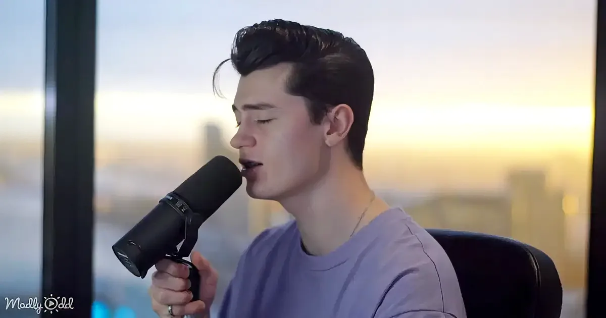 A person sings into a microphone, wearing a purple shirt, with a cityscape and sunset in the background.