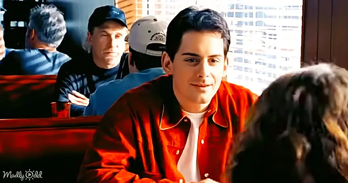 A man in a red jacket smiles at someone across a diner booth, with other patrons in the background.