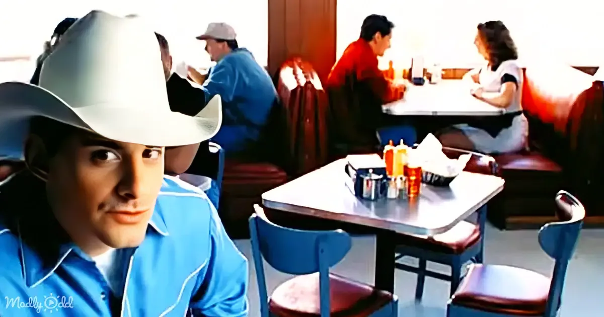 A man in a white cowboy hat and blue shirt sits in a diner, looking directly at the camera.
