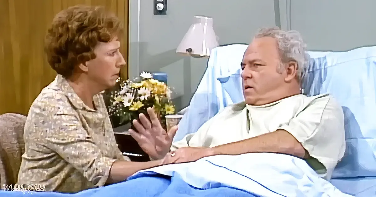 A woman in a floral shirt talks to a man in a hospital bed, with a bouquet of flowers nearby.
