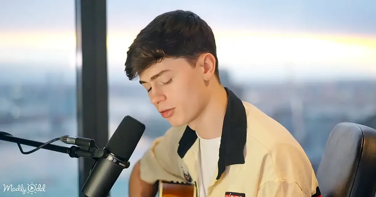 A person in a light shirt sits by a microphone, playing guitar, with a cityscape in the background.