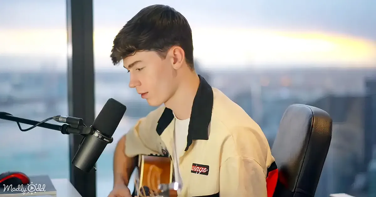 A young man plays guitar and sings into a microphone, wearing a light-colored jacket with a "Wings" patch, against a...