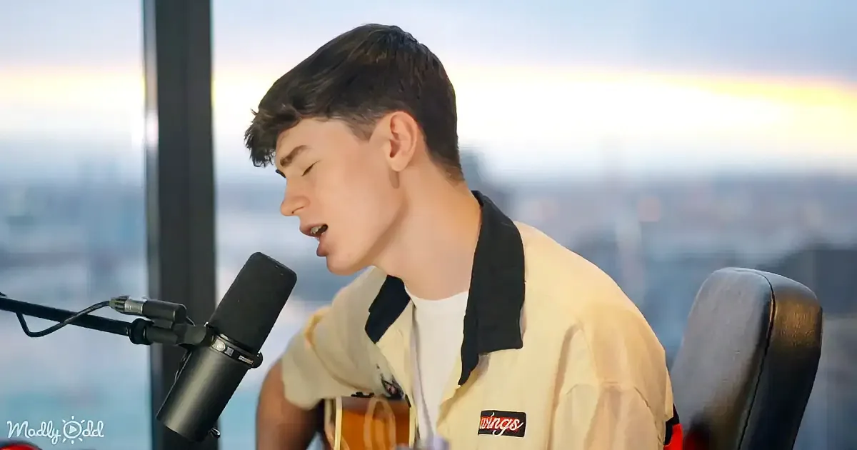 A young man sings into a microphone, holding a guitar, with a cityscape in the background.