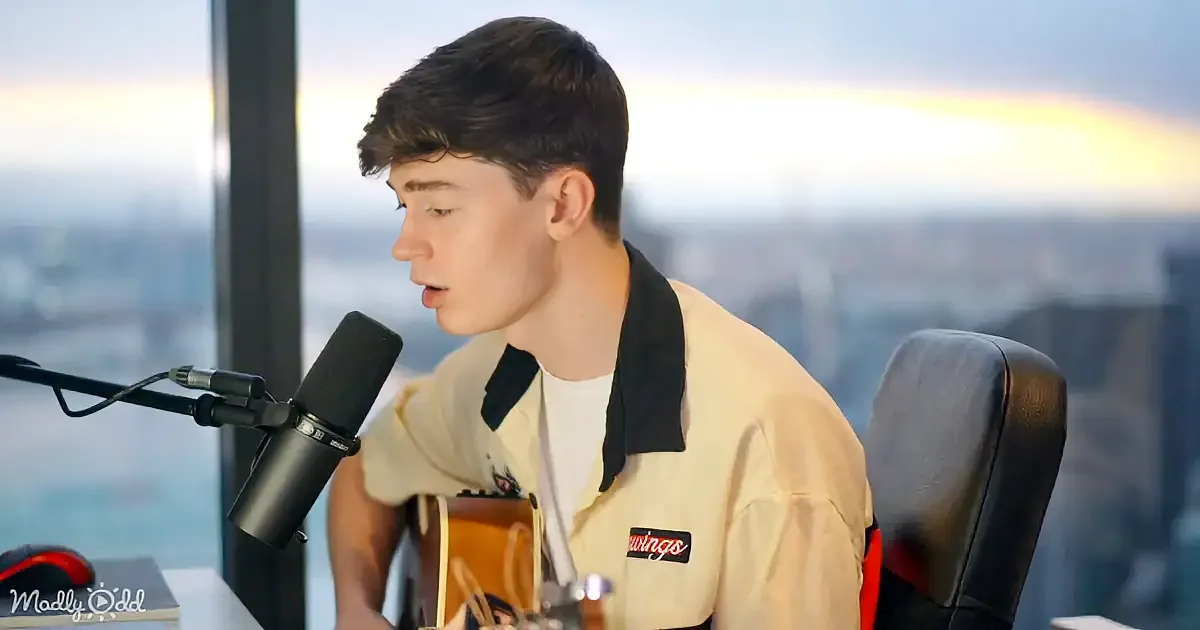 A young man sings into a microphone while playing guitar, wearing a light-colored jacket with a "wings" patch, against a...