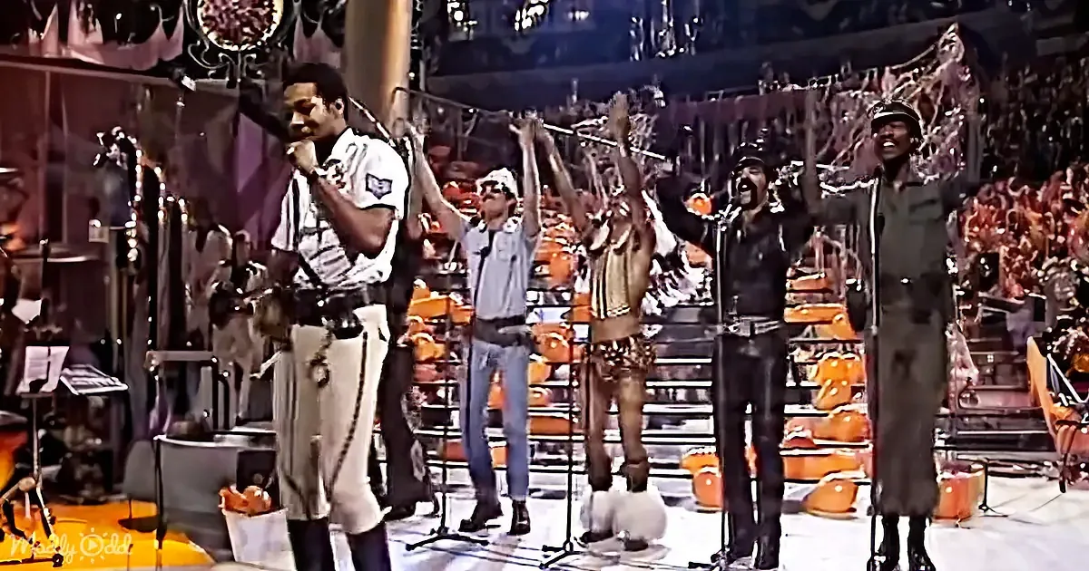 A group of performers in military-style uniforms sing on stage, surrounded by musicians and a large audience.