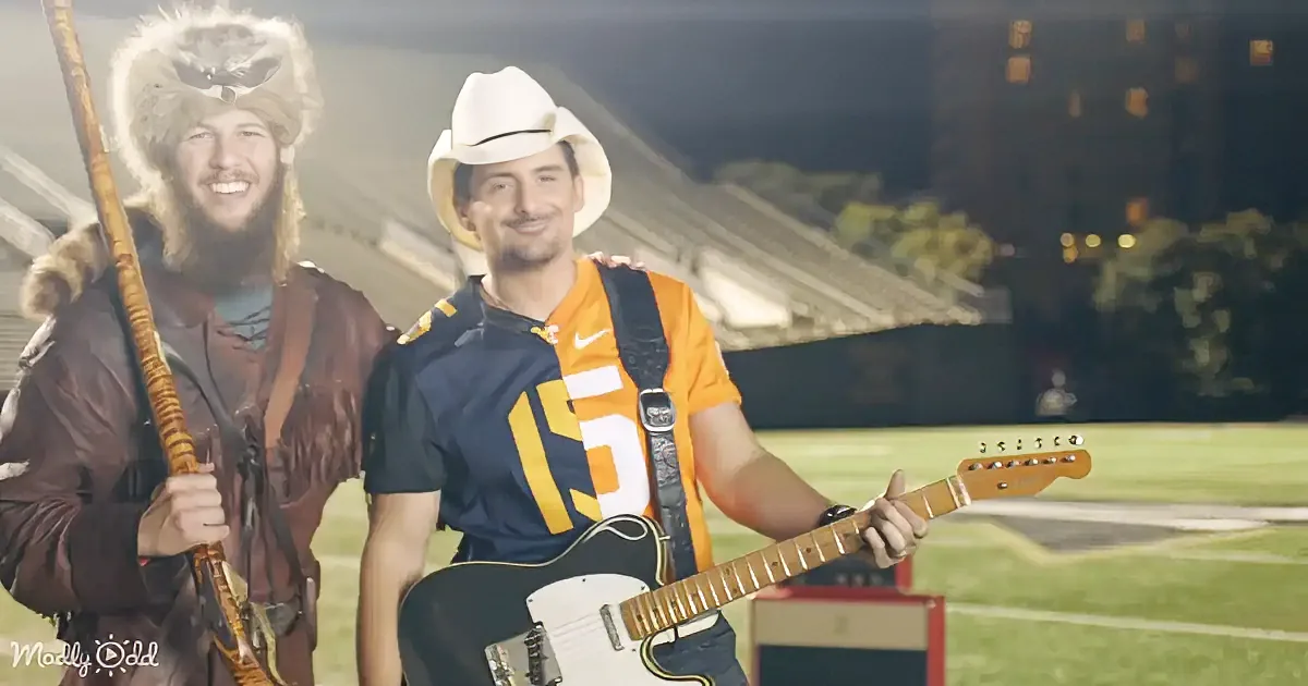 A man in a Tennessee football jersey and cowboy hat holds a guitar on a field.