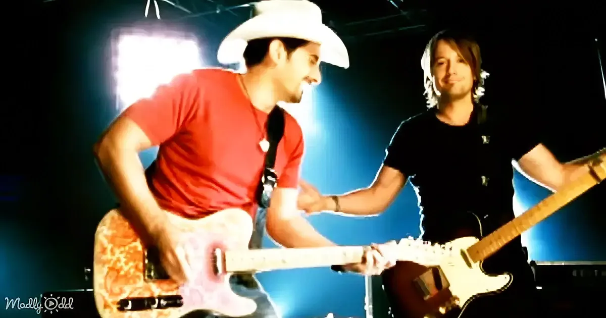 Two musicians in red and black shirts shake hands while playing guitars on stage.