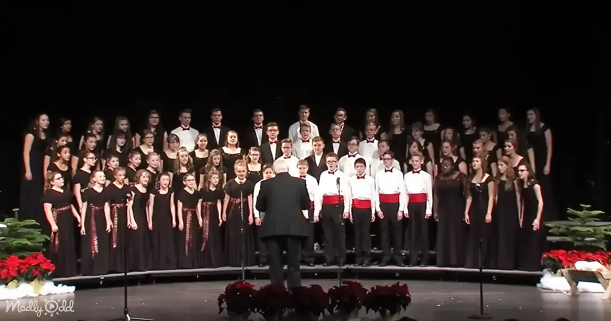 A conductor leads a choir in formal attire on stage with festive decorations.