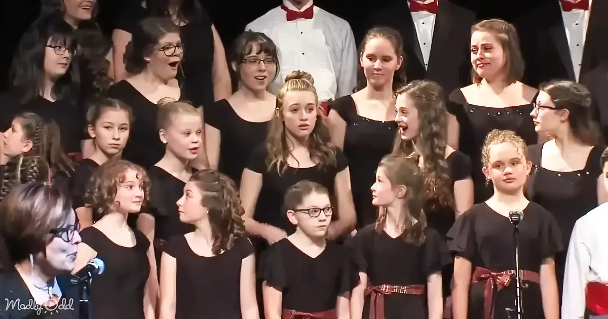 A group of young girls in black dresses sing together, some wearing glasses, with a few adults in formal attire behind them.