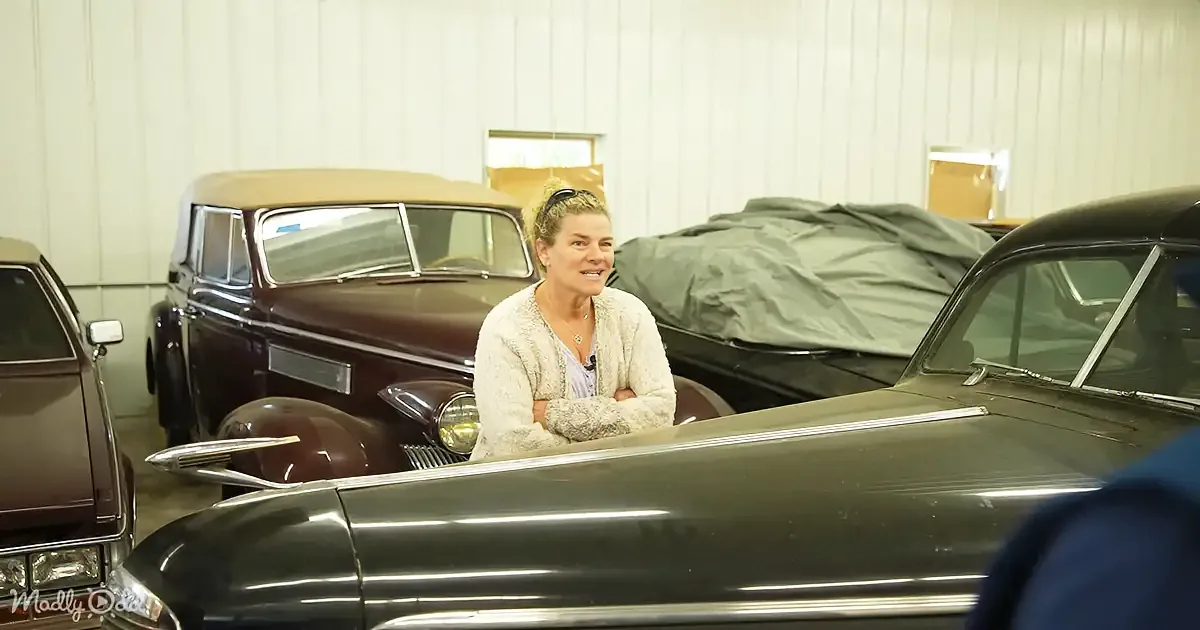 A woman stands smiling in a garage surrounded by vintage cars, including a maroon convertible and a dark-colored sedan.