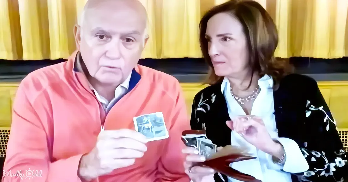 A man in a red sweater holds a card, while a woman in a black and white outfit looks on, both seated in front of yellow...