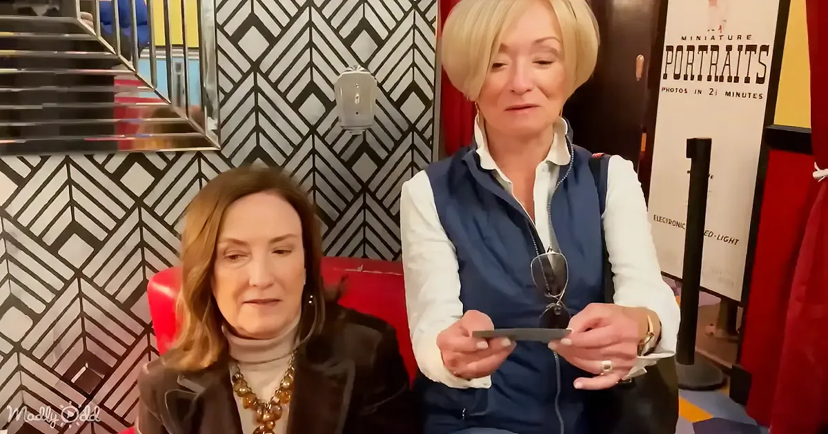 Two women sit in a room with a geometric patterned wall, one holding a small object.