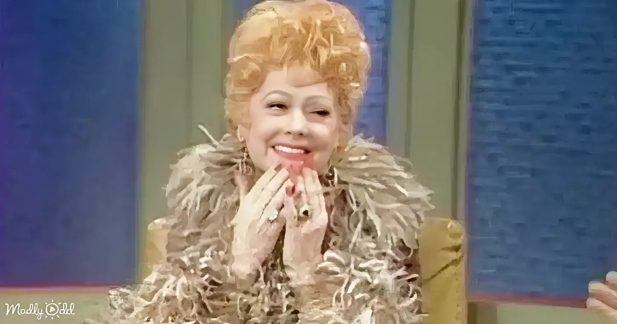 A person with curly blonde hair and a feathered outfit is smiling and holding their hands near their face.