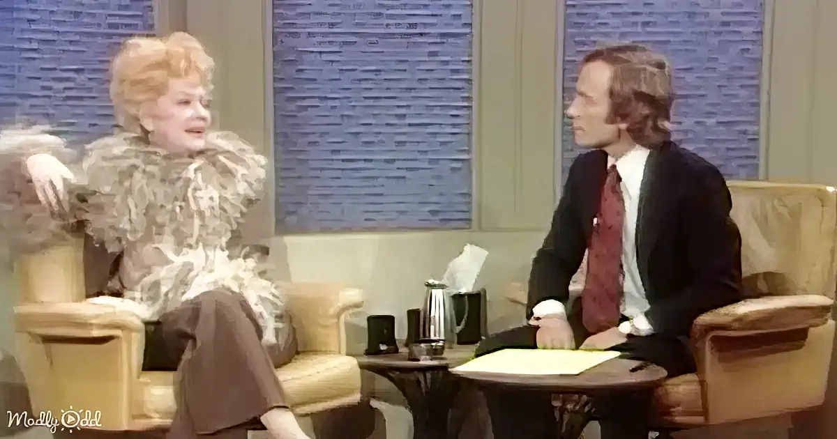 A woman in a fur coat gestures animatedly while seated, with a man in a suit listening attentively.