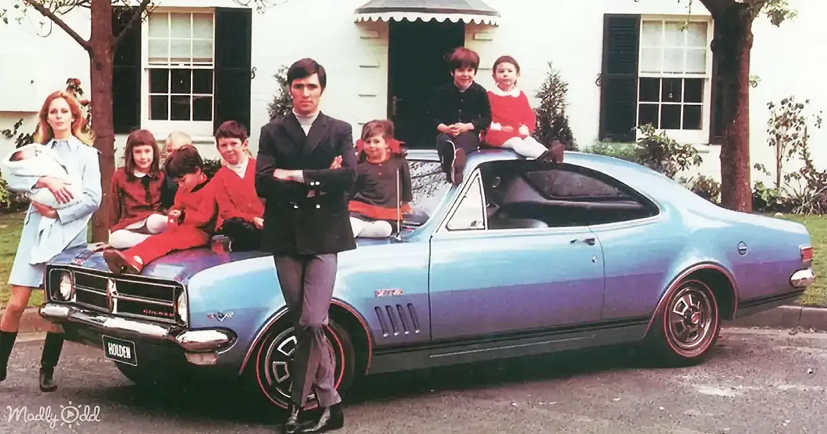 A man stands beside a blue Holden car, surrounded by children and a woman holding a baby, in front of a white house.