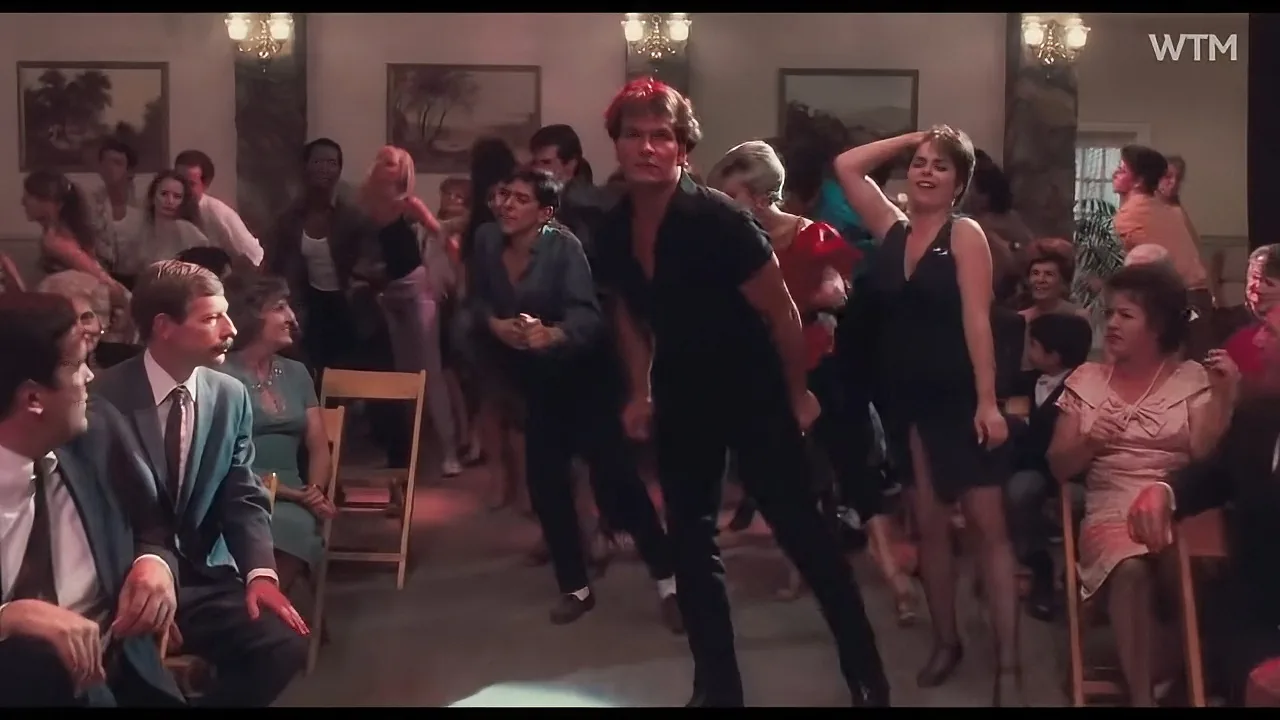 A man in a black outfit dances energetically in a crowded room with people watching.