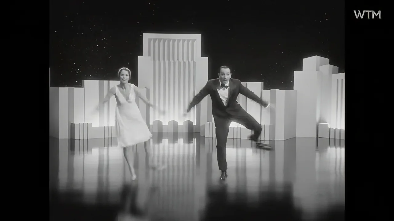 A man in a tuxedo and a woman in a dress are dancing in front of a cityscape backdrop.