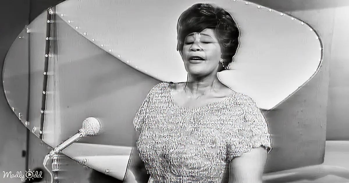A woman in a textured dress sings into a microphone, with a large decorative mirror behind her.