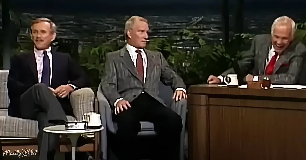 Three men in suits sit on a talk show set, one laughing, another looking serious, and the third appearing relaxed.