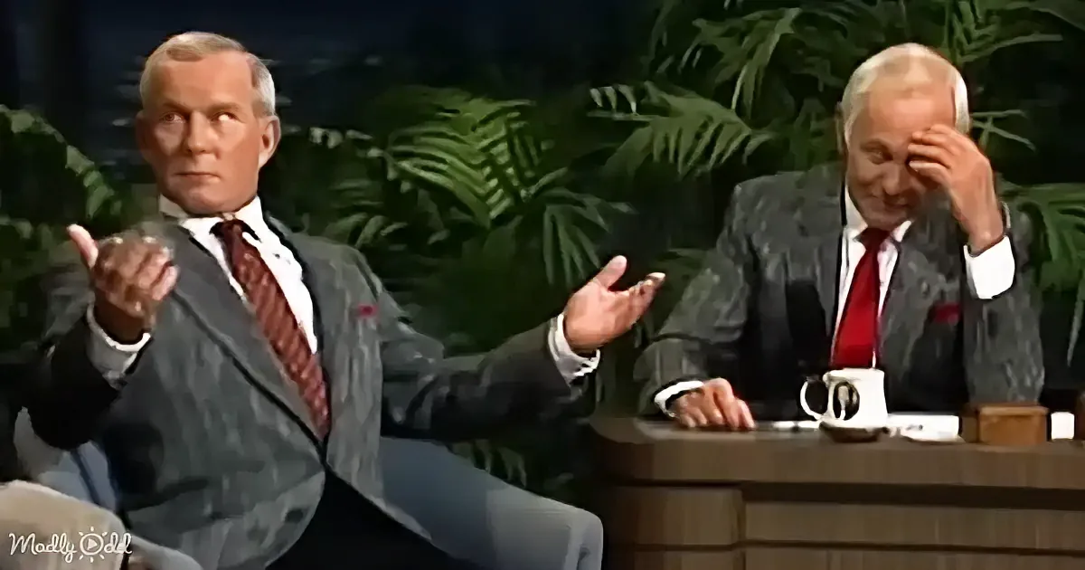 Two men in suits, one gesturing, the other appearing distressed, seated in a studio setting with plants in the background.