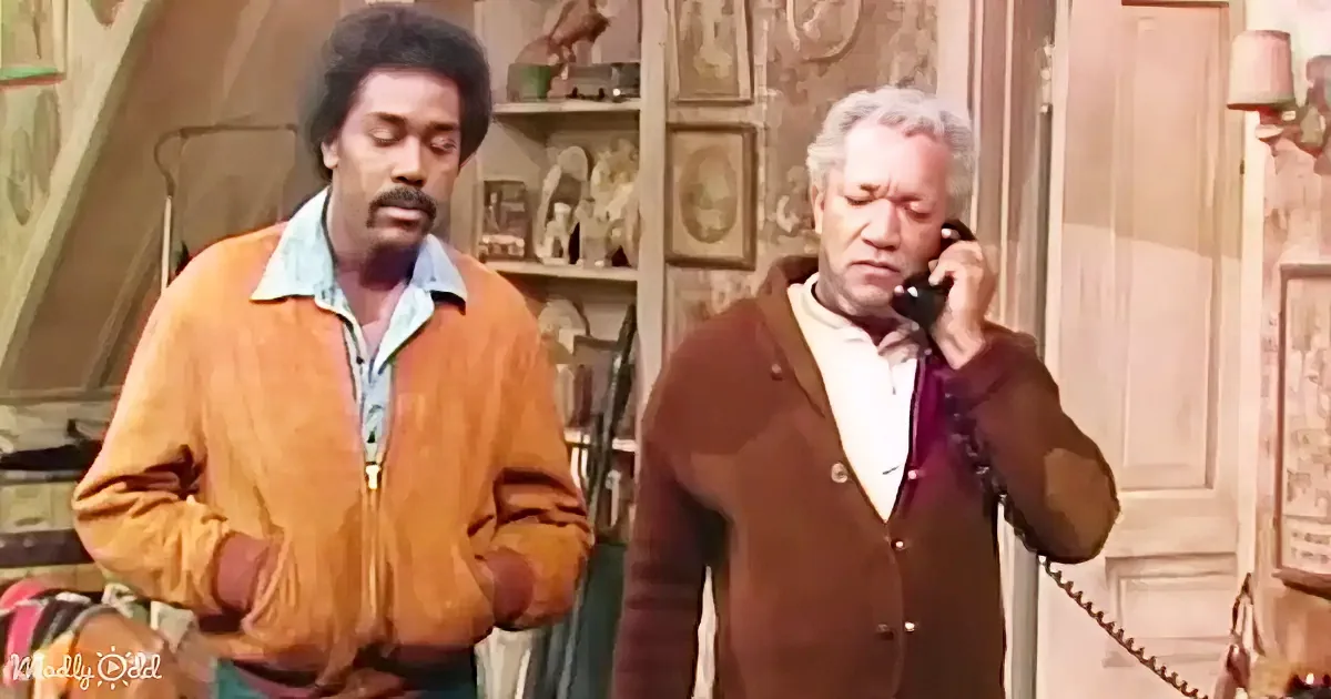 A man in an orange jacket stands beside another man talking on a phone in a room with shelves and artwork.