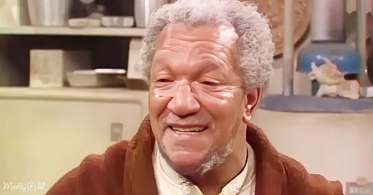 An older man with white hair and a brown jacket smiles in a kitchen setting.
