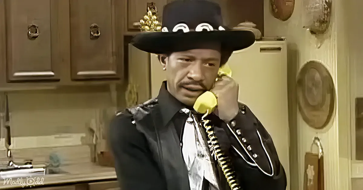 A man in a black hat and jacket is talking on a yellow phone in a kitchen.