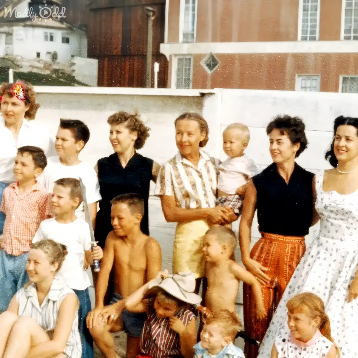 A group of women and children, dressed in vintage attire, pose together outdoors, with a building and wall in the background.