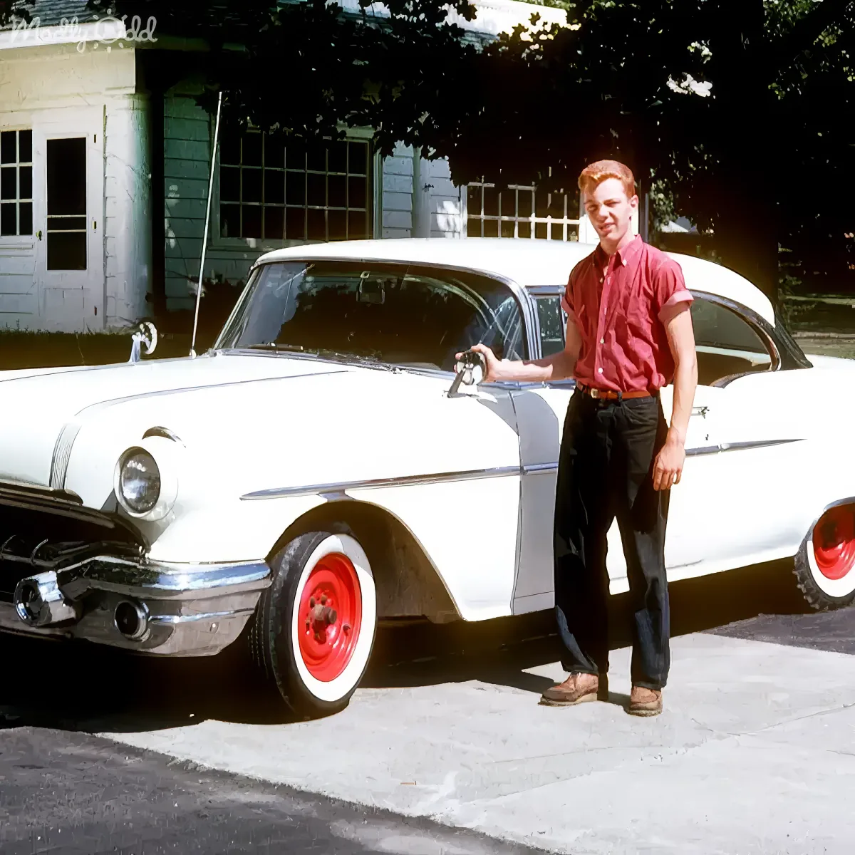 A man in a red shirt stands beside a white vintage car with red wheels, smiling.