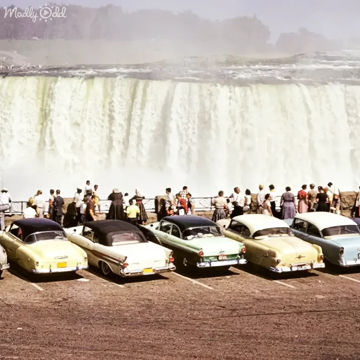 People gather around vintage cars in a parking lot, overlooking a powerful waterfall.