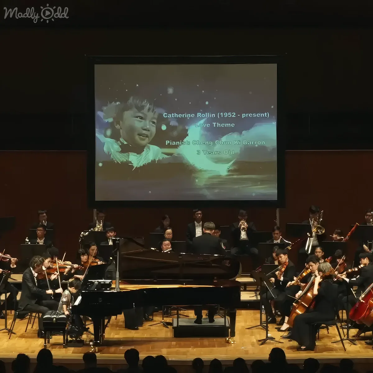 A young child is projected on a screen above an orchestra performing a live theme.