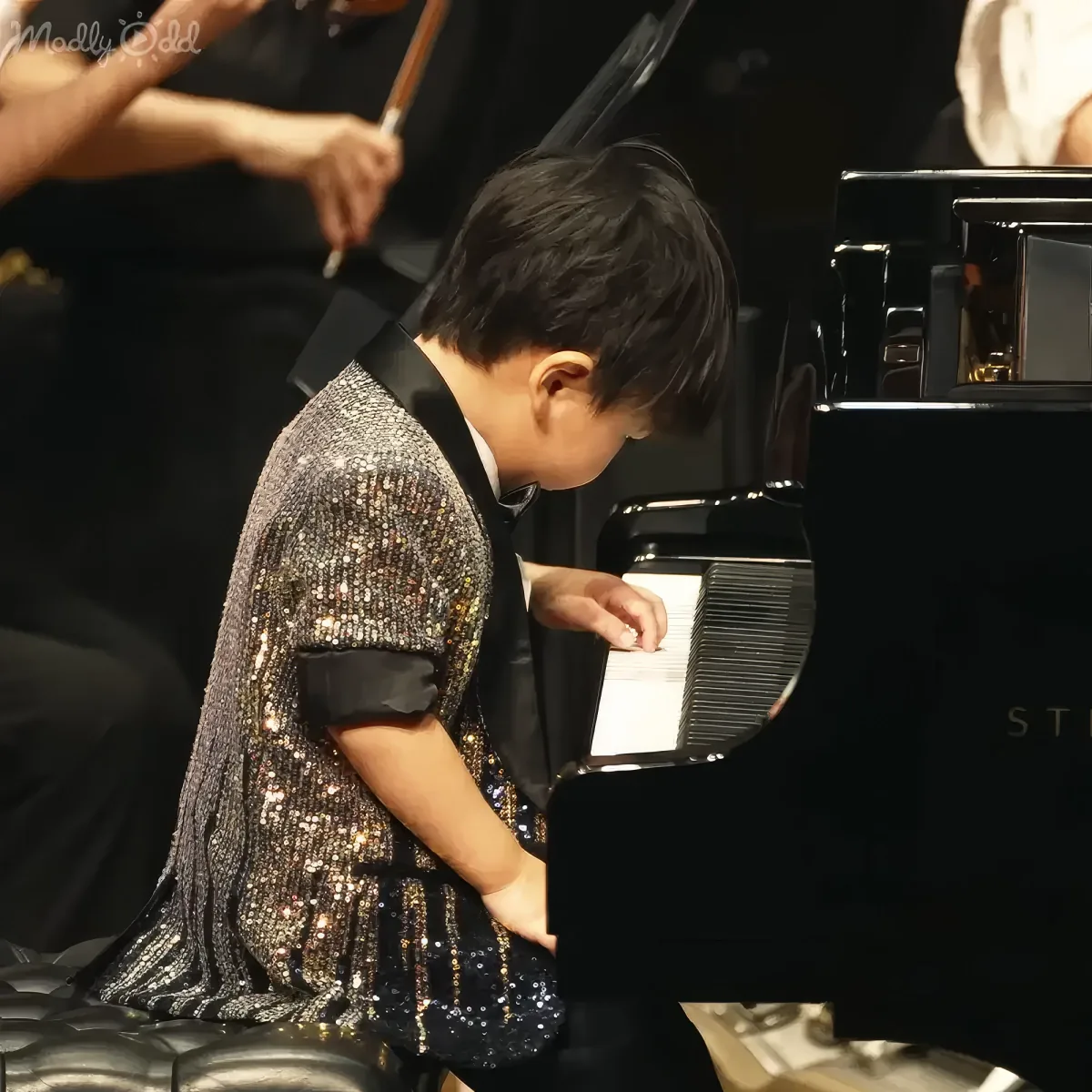 A young child in a sparkly dress plays a grand piano, surrounded by musicians.
