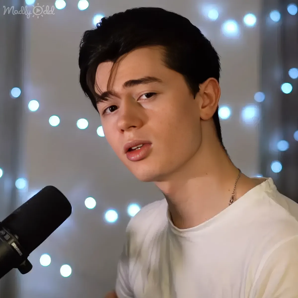 A person wearing a white shirt and a necklace, speaking into a microphone with string lights in the background.