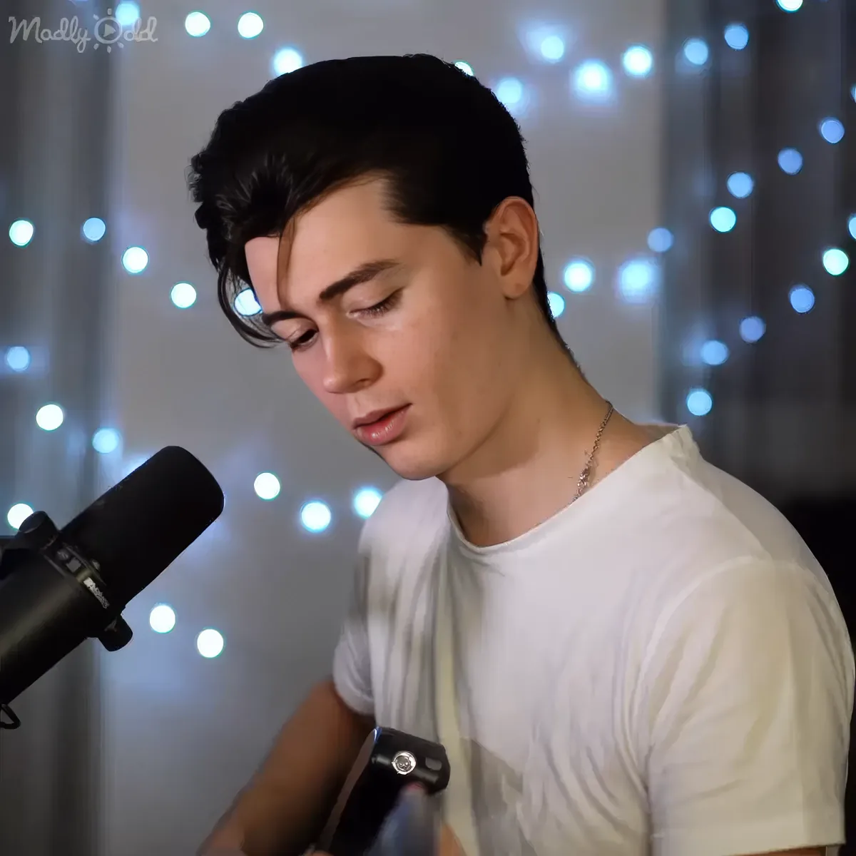 A person in a white shirt plays guitar in front of a backdrop of twinkling lights.