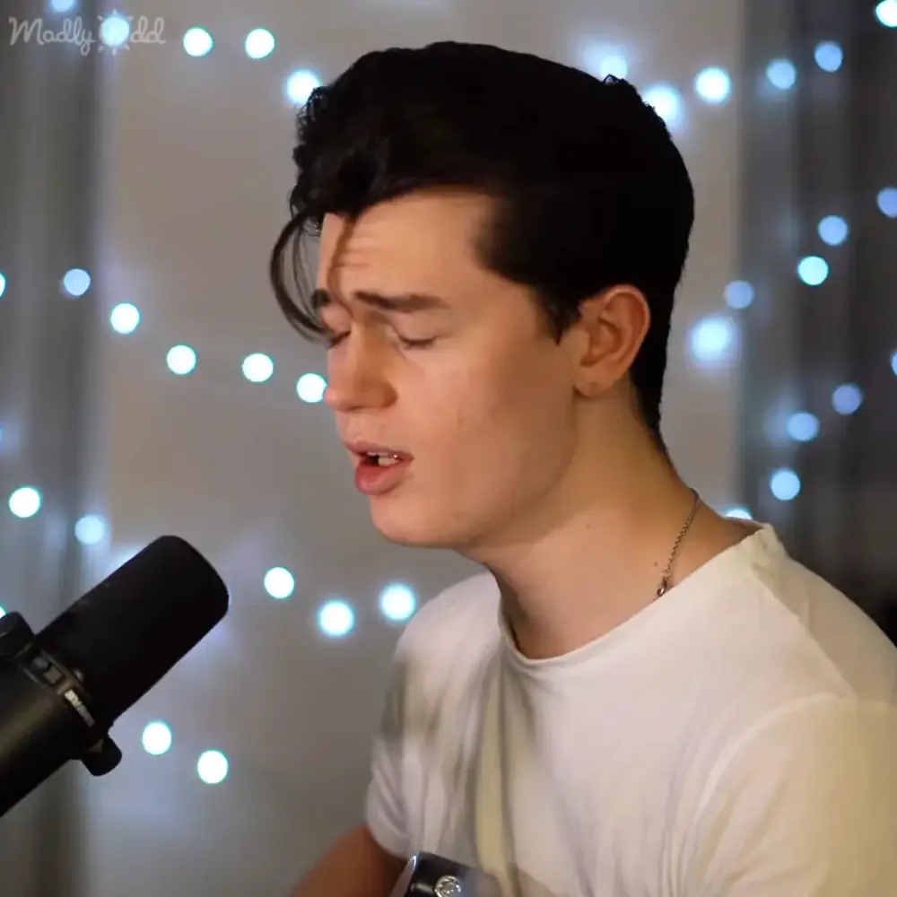 A person in a white shirt sings into a microphone, with string lights in the background.